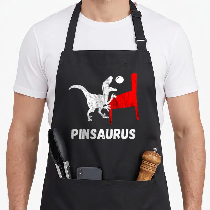 Pinball dinosaur apron - White dinosaur and red pinball machine graphic with centered PINSAURUS text — shown on a black cotton canvas apron against a white background