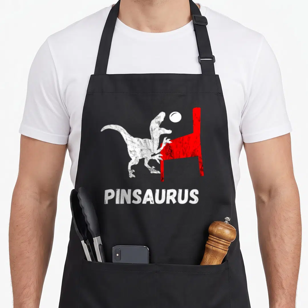 Pinball dinosaur apron - White dinosaur and red pinball machine graphic with centered PINSAURUS text — shown on a black cotton canvas apron against a white background