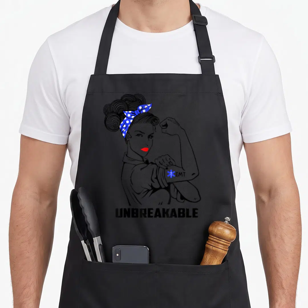 Unbreakable emt apron - Cartoon woman flexing arm with EMT symbol and UNBREAKABLE text in black white blue red — shown on a black cotton canvas apron against a white background