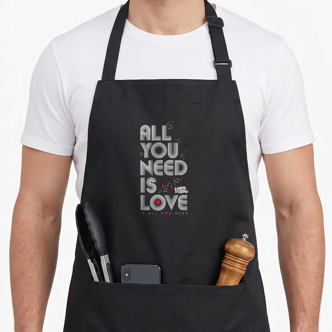 Lyric band lover apron - Stacked white text All You Need Is Love with red hearts and musical notes — shown on a black cotton canvas apron against a white background