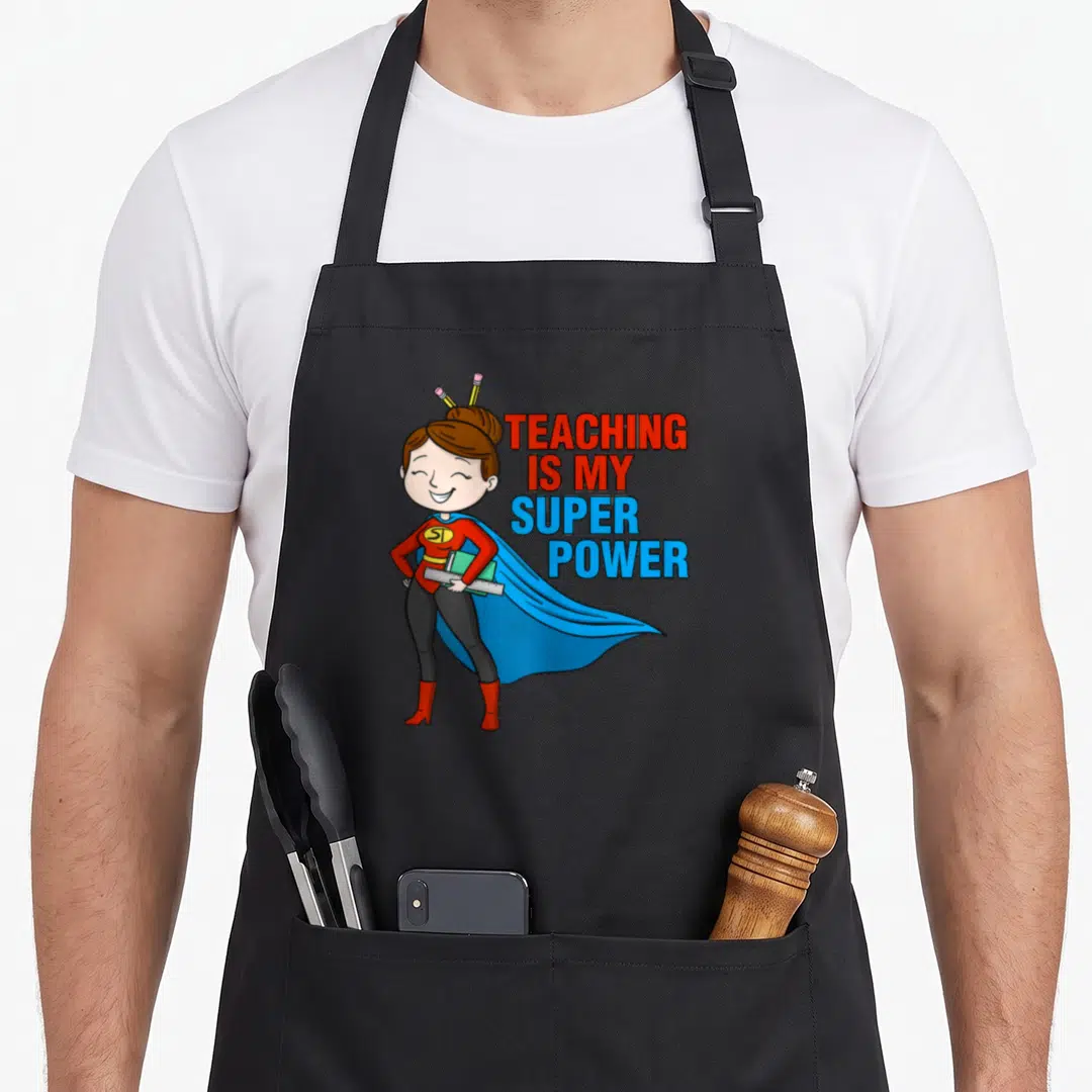 Teaching kindergarten teacher apron - Cartoon superhero woman holding green book and ruler with pencils in hair — shown on a black cotton canvas apron against a white background