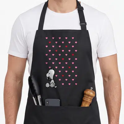 Cute valentine apron - White cartoon dog sitting among scattered pink and red hearts — shown on a black cotton canvas apron against a white background