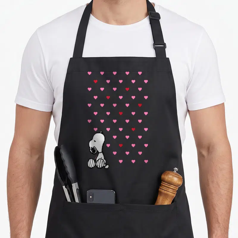 Cute valentine apron - White cartoon dog sitting among scattered pink and red hearts — shown on a black cotton canvas apron against a white background