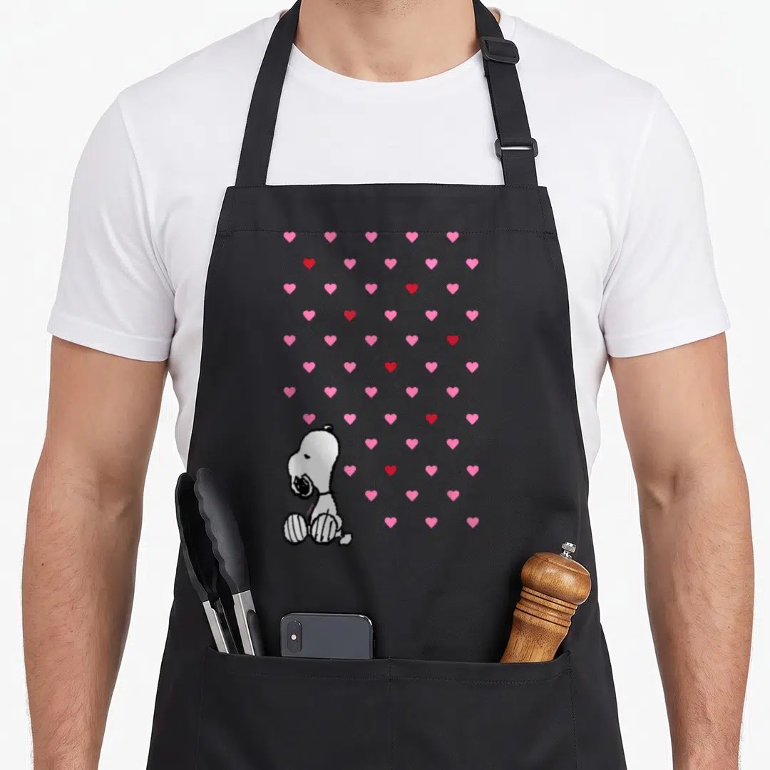 Cute valentine apron - White cartoon dog sitting among scattered pink and red hearts — shown on a black cotton canvas apron against a white background