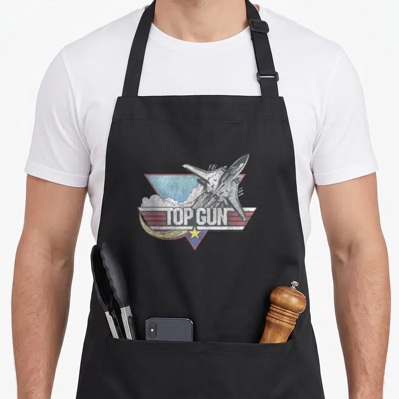 Funny jet gun apron - Distressed TOP GUN fighter jet graphic with yellow star and red stripes on black apron — shown on a black cotton canvas apron against a white background