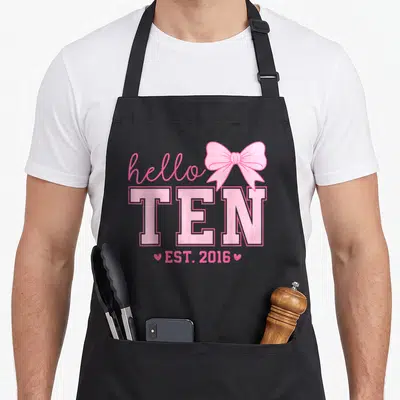 Hello bow apron - Pink hello TEN EST 2016 text with bow and two hearts — shown on a black cotton canvas apron against a white background