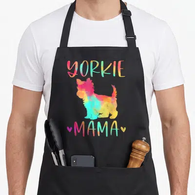 Mama colorful yorkie apron - Rainbow watercolor Yorkshire Terrier silhouette with YORKIE MAMA text and two hearts — shown on a black cotton canvas apron against a white background
