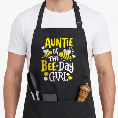 Auntie birthday bee aprons - Stacked AUNTIE OF THE BEE-Day GIRL text with two bees and flowers — shown on a black cotton canvas apron against a white background