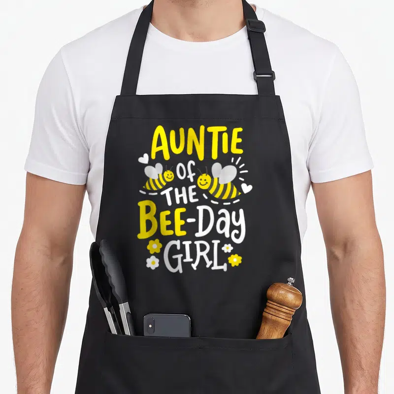 Auntie birthday bee aprons - Stacked AUNTIE OF THE BEE-Day GIRL text with two bees and flowers — shown on a black cotton canvas apron against a white background