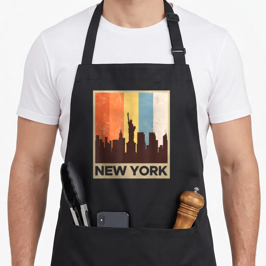70s new york apron - Retro NYC skyline silhouette with Statue of Liberty on distressed red orange yellow blue and cream — shown on a black cotton canvas apron against a white background
