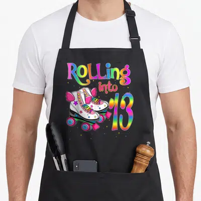 Cute rolling turning apron - Rolling into 13 text with white roller skates, pink wings, rainbow letters, and stars — shown on a black cotton canvas apron against a white background