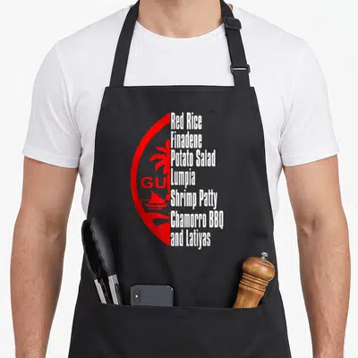Fiesta guam aprons - Red half circle with palm tree and boat next to stacked white text listing Guam dishes like Red — shown on a black cotton canvas apron against a white background