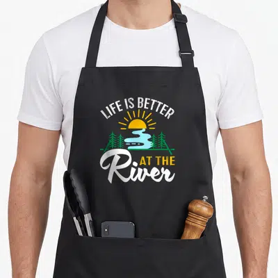 Better tree apron - Cartoon sun river and trees with Life Is Better At The River text in white yellow green and blue — shown on a black cotton canvas apron against a white background