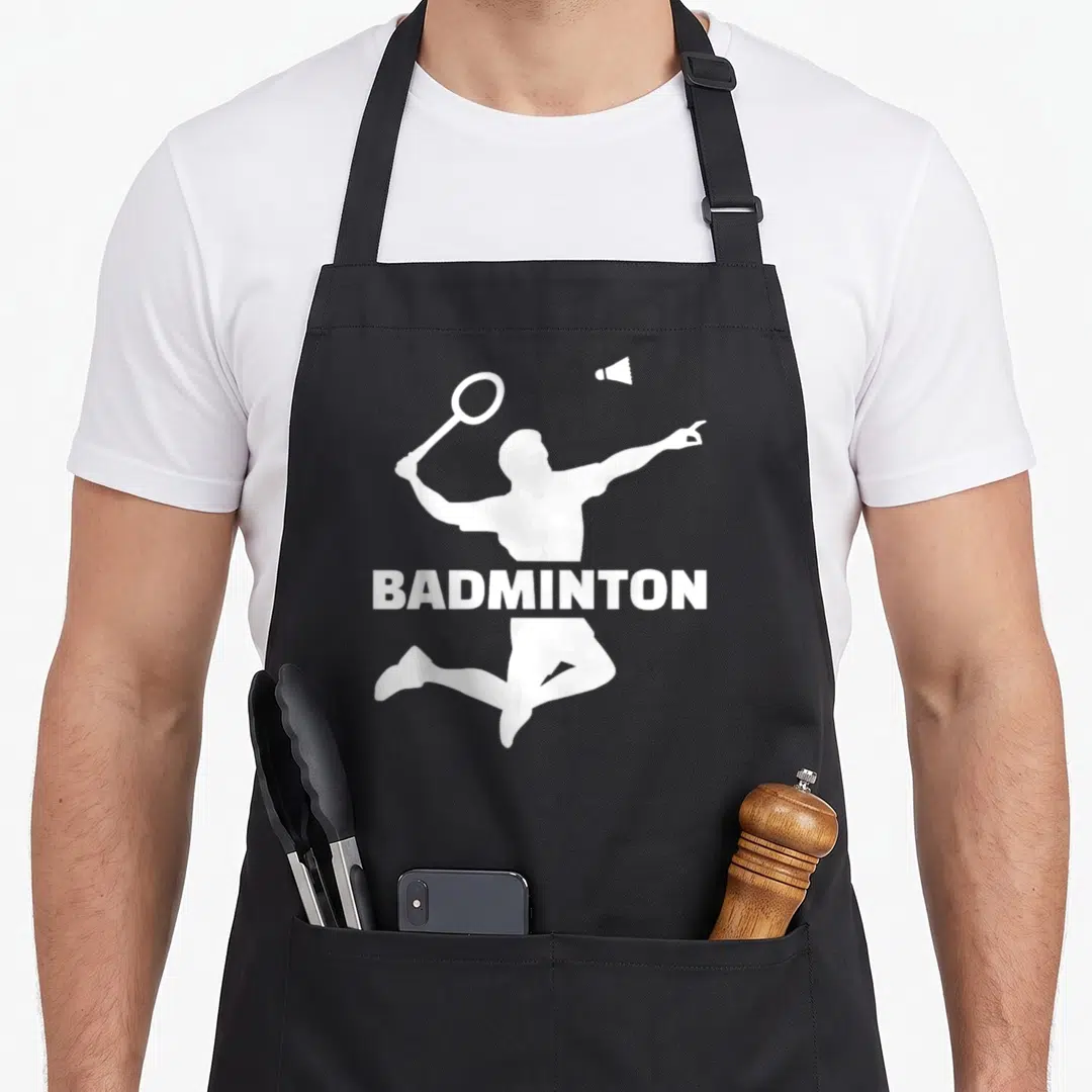 Cute player badminton apron - White jumping badminton player silhouette with racket and shuttlecock, text BADMINTON — shown on a black cotton canvas apron against a white background