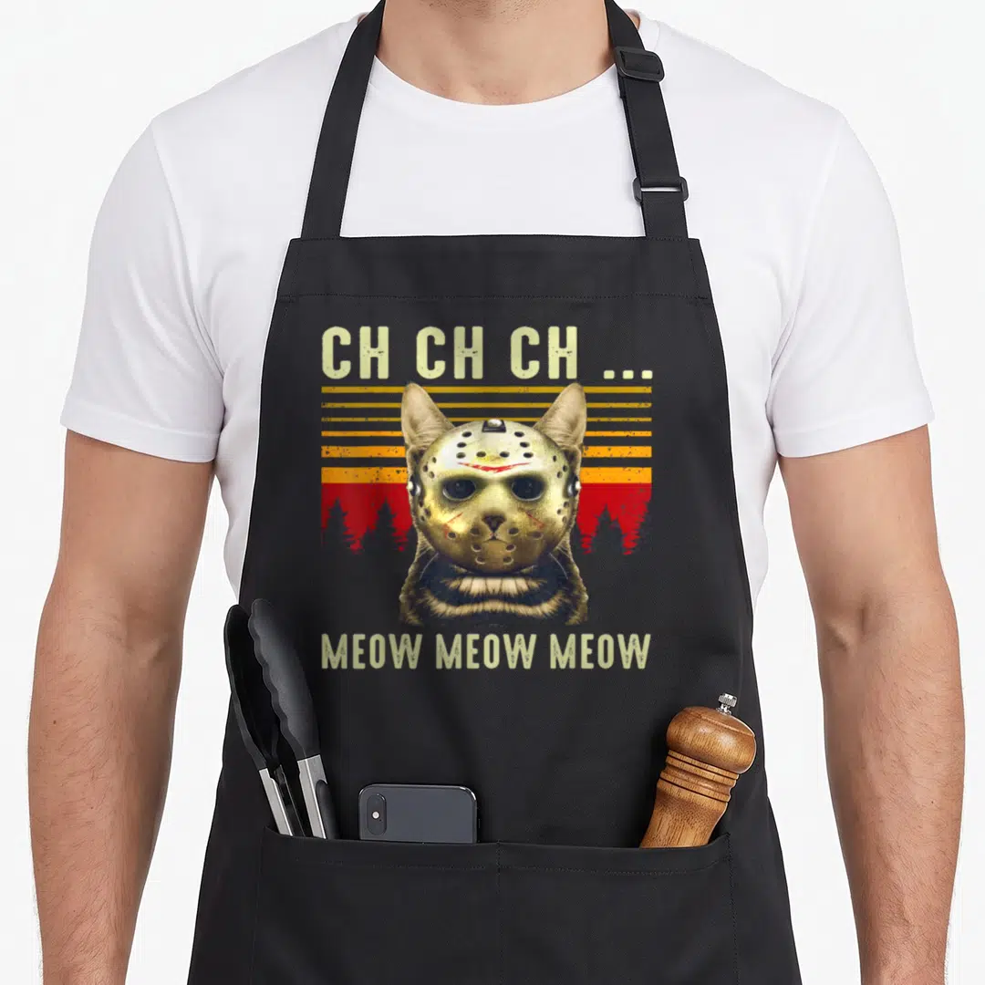 Scary cat apron - Cat wearing hockey mask with CH CH CH and MEOW text on retro sunset background — shown on a black cotton canvas apron against a white background