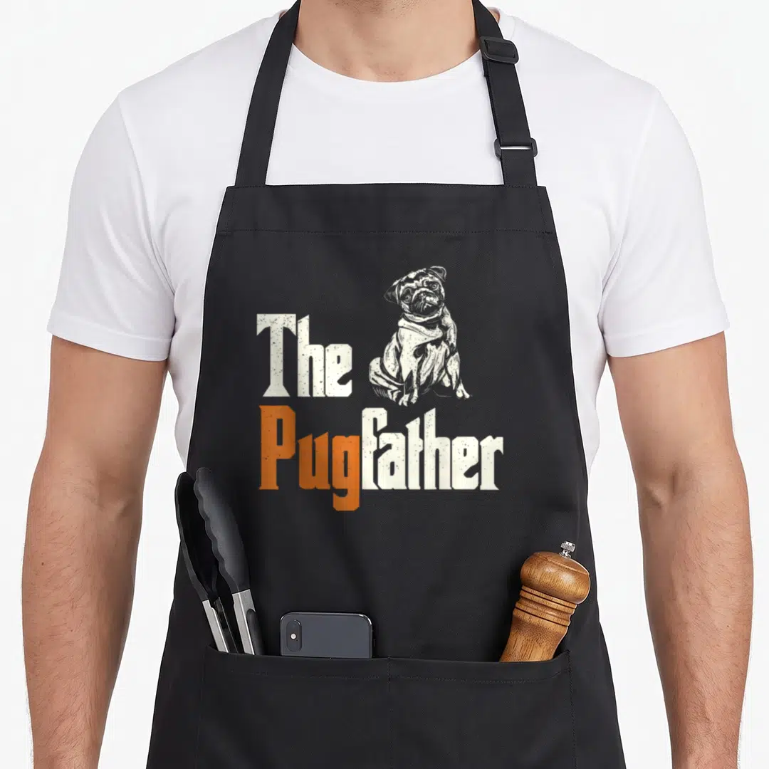 Pugfather dog apron - Sitting pug illustration next to distressed white and orange text reading The PugFather — shown on a black cotton canvas apron against a white background