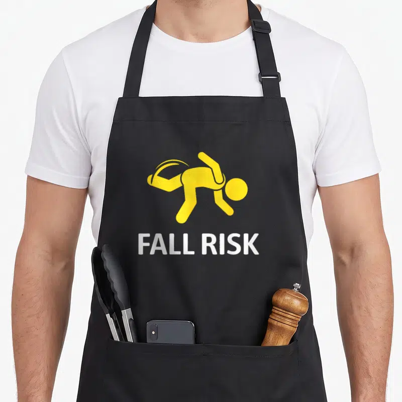Issue fall apron - Yellow stick figure falling over white FALL RISK text on black background — shown on a black cotton canvas apron against a white background