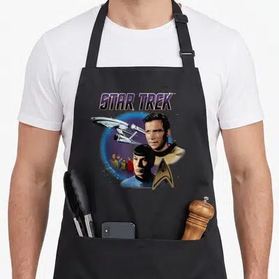 Original star apron - STAR TREK logo above Captain Kirk, USS Enterprise, and crew against blue purple starfield — shown on a black cotton canvas apron against a white background