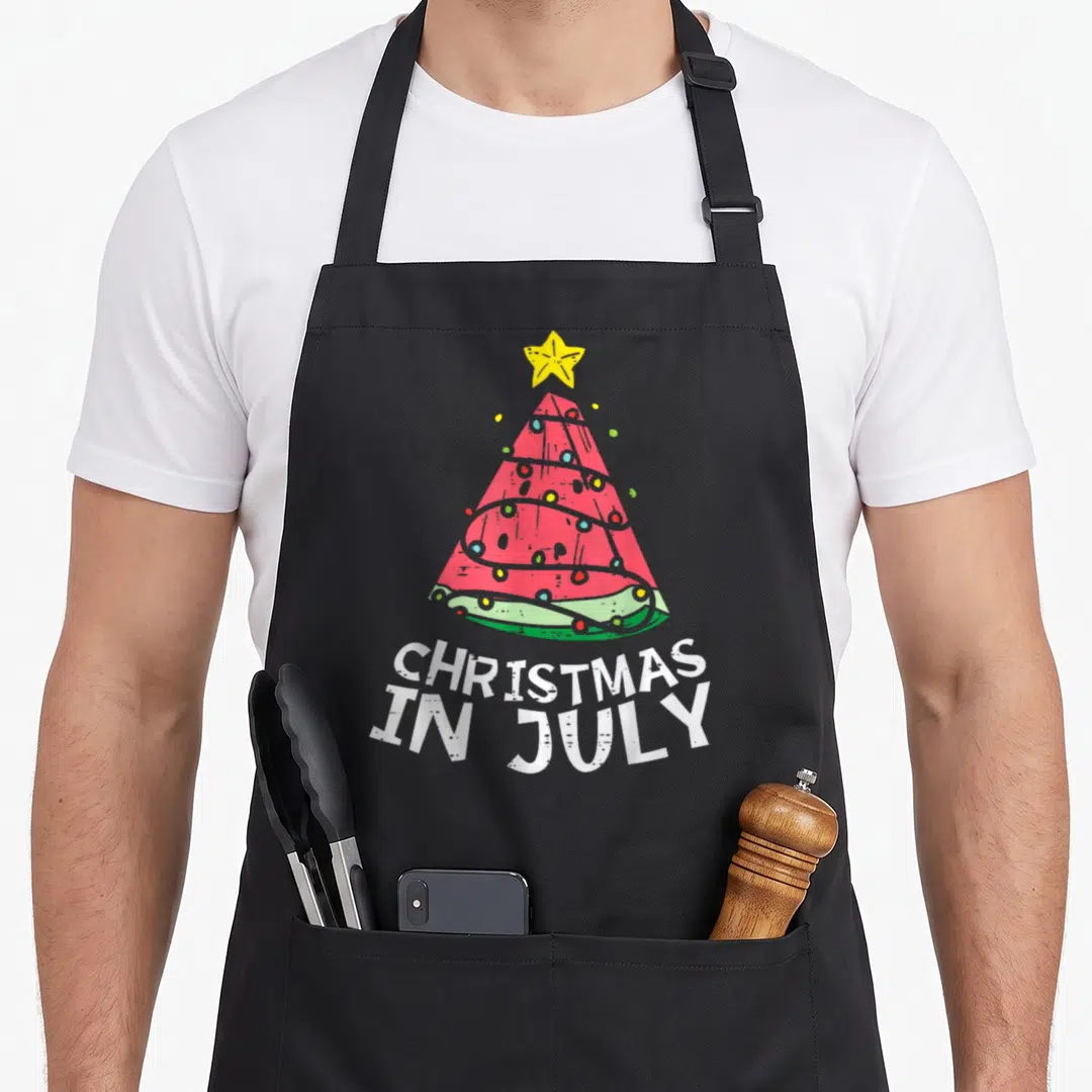 Christmas watermelon apron - Red watermelon slice shaped as a tree with lights and white text reading CHRISTMAS IN JULY — shown on a black cotton canvas apron against a white background
