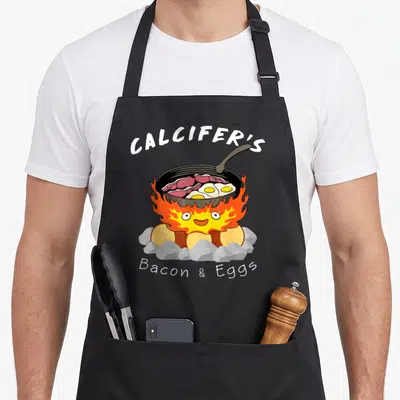 Food bacon apron - Cartoon fire demon cooking bacon and eggs in pan with arched CALCIFER'S text — shown on a black cotton canvas apron against a white background