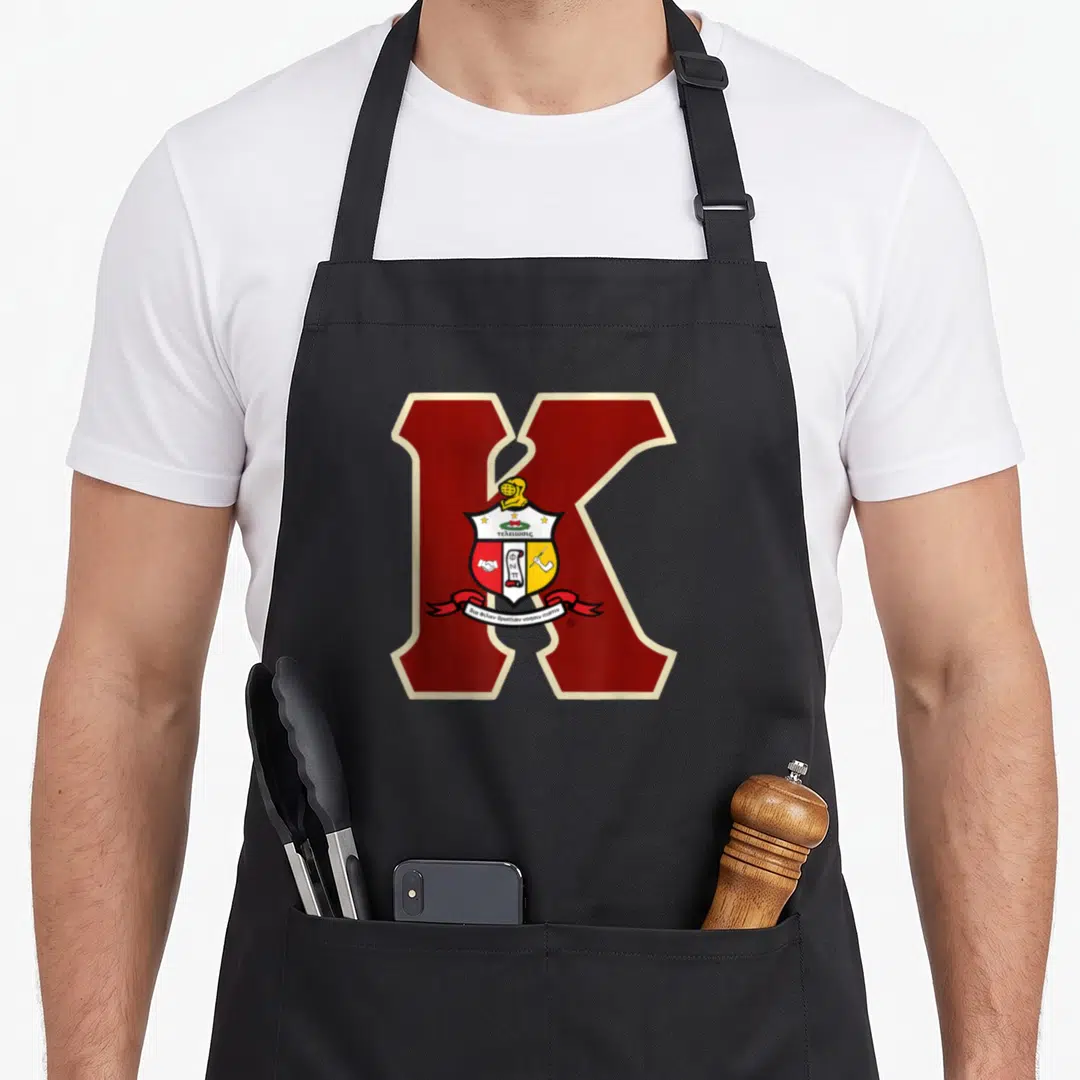 Cute psi alpha apron - Maroon K with crest and Greek text τελειωσις on black apron — shown on a black cotton canvas apron against a white background