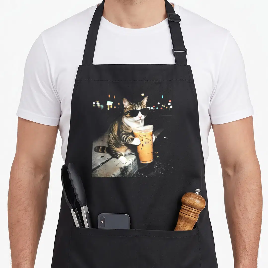 Drinking cat apron - Tabby cat wearing black sunglasses sipping orange iced drink from a plastic cup on a grey stone — shown on a black cotton canvas apron against a white background