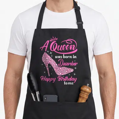 Funny december birthday apron - Pink high heel shoe with snowflake pattern and crown above text A Queen was born — shown on a black cotton canvas apron against a white background