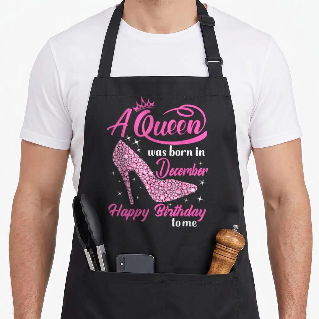 Funny december birthday apron - Pink high heel shoe with snowflake pattern and crown above text A Queen was born — shown on a black cotton canvas apron against a white background