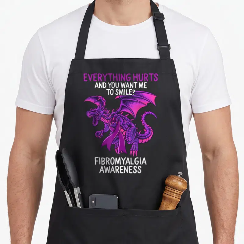 Fibromyalgia dragon apron - Purple dragon with ribbon and white text reading Everything Hurts Fibromyalgia Awareness — shown on a black cotton canvas apron against a white background