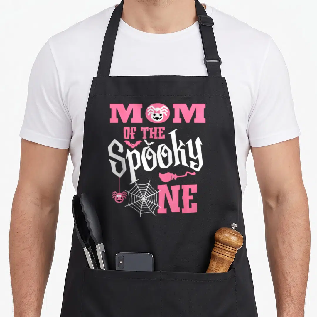 1st birthday funny halloween apron - Pink and white MOM OF THE Spooky ONE text with spider in O, bats, web, and worm — shown on a black cotton canvas apron against a white background
