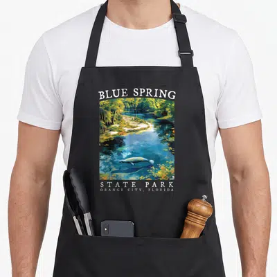 Manatee spring apron - Realistic manatee swimming in blue water under arched BLUE SPRING text — shown on a black cotton canvas apron against a white background