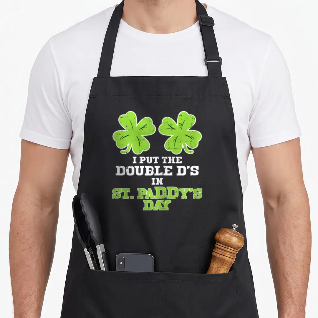 Paddy double apron - Two green shamrocks above stacked text reading I put the double d's in St Paddys Day — shown on a black cotton canvas apron against a white background