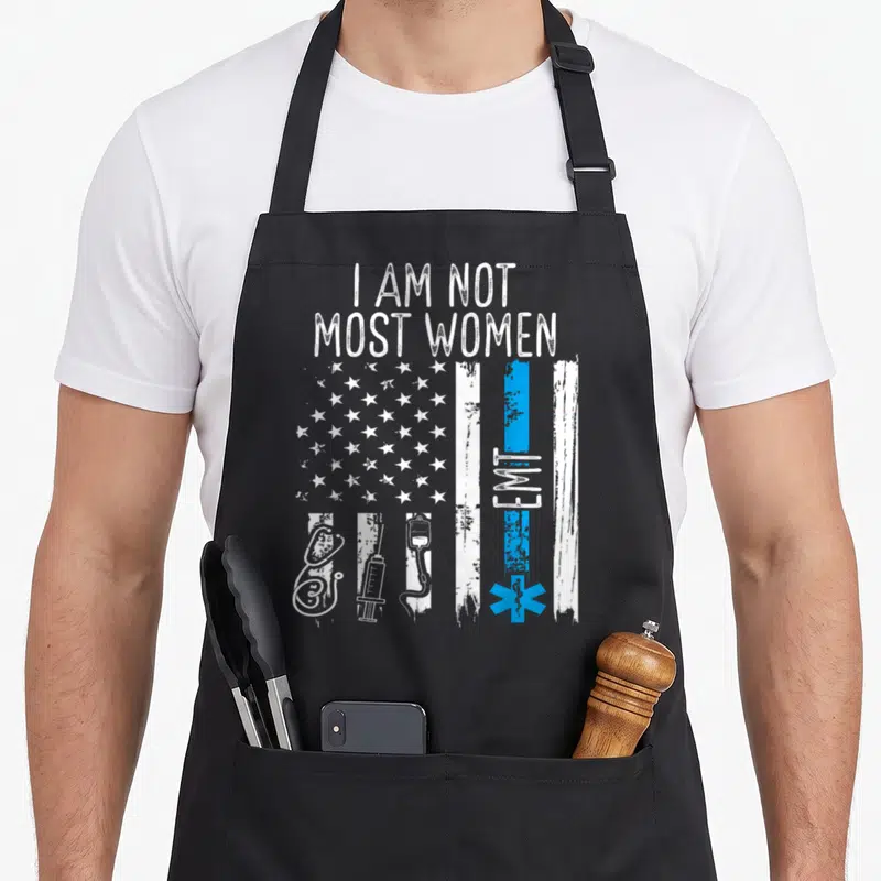 Emt healthcare apron - Distressed flag with I AM NOT MOST WOMEN text and medical equipment stripes in white and blue — shown on a black cotton canvas apron against a white background