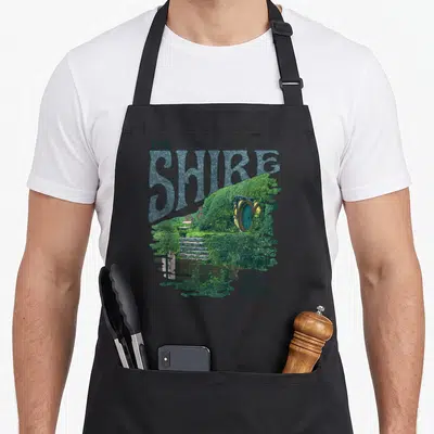 Lotr rings apron - Retro print of The Shire hobbit hole with green hill and blue-grey lettering — shown on a black cotton canvas apron against a white background