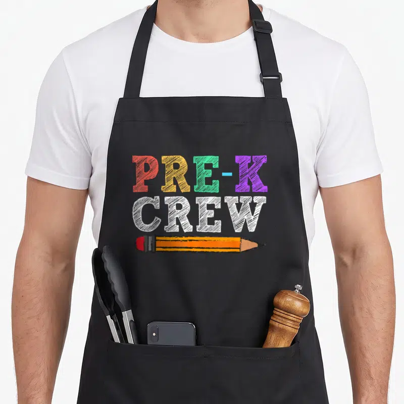 Pre crew teacher apron - PRE-K CREW text in red yellow green purple white with horizontal pencil below — shown on a black cotton canvas apron against a white background