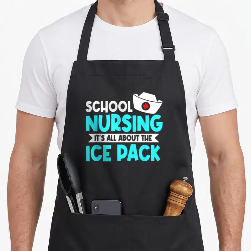 Health nursing student apron - Stacked text SCHOOL NURSING IT'S ALL ABOUT THE ICE PACK with nurse hat and red cross — shown on a black cotton canvas apron against a white background