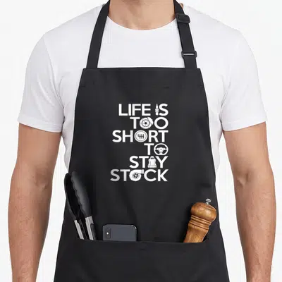 Classical stock aprons - White stacked text LIFE IS TOO SHORT TO STAY STOCK with car part icons replacing letters — shown on a black cotton canvas apron against a white background