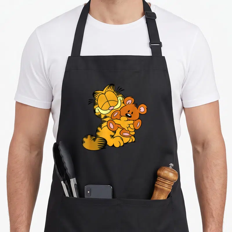 Hugging cat apron - Orange cartoon cat sitting and hugging a brown teddy bear in a wholesome embrace — shown on a black cotton canvas apron against a white background