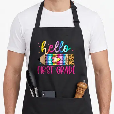 Grade tie-dye apron - Hello First Grade pencil with heart, leopard eraser, tie-dye colors, pink dots — shown on a black cotton canvas apron against a white background
