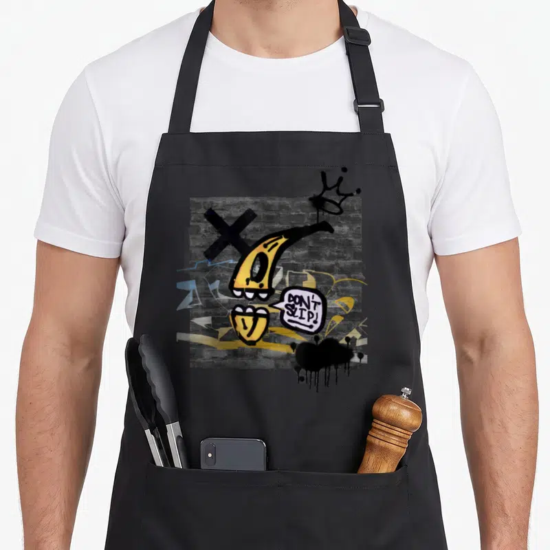Graffiti slip on apron - Yellow cartoon banana peel with DON'T SLIP speech bubble on grey brick wall — shown on a black cotton canvas apron against a white background