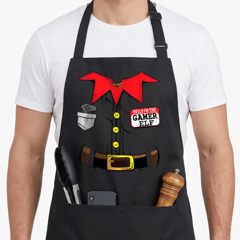 Stocking christmas elf apron - Cartoon elf torso with controller pocket and HELLO I'M THE GAMER ELF name tag — shown on a black cotton canvas apron against a white background
