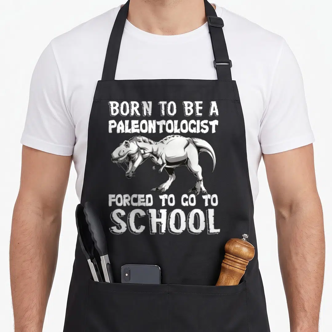 Dinausor forced school apron - Cartoon T-Rex with white text Born to be a paleontologist forced to go to school — shown on a black cotton canvas apron against a white background