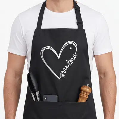 Christmas grandma in apron - White line-art heart with grandma text forming the right edge and a tiny inner heart — shown on a black cotton canvas apron against a white background