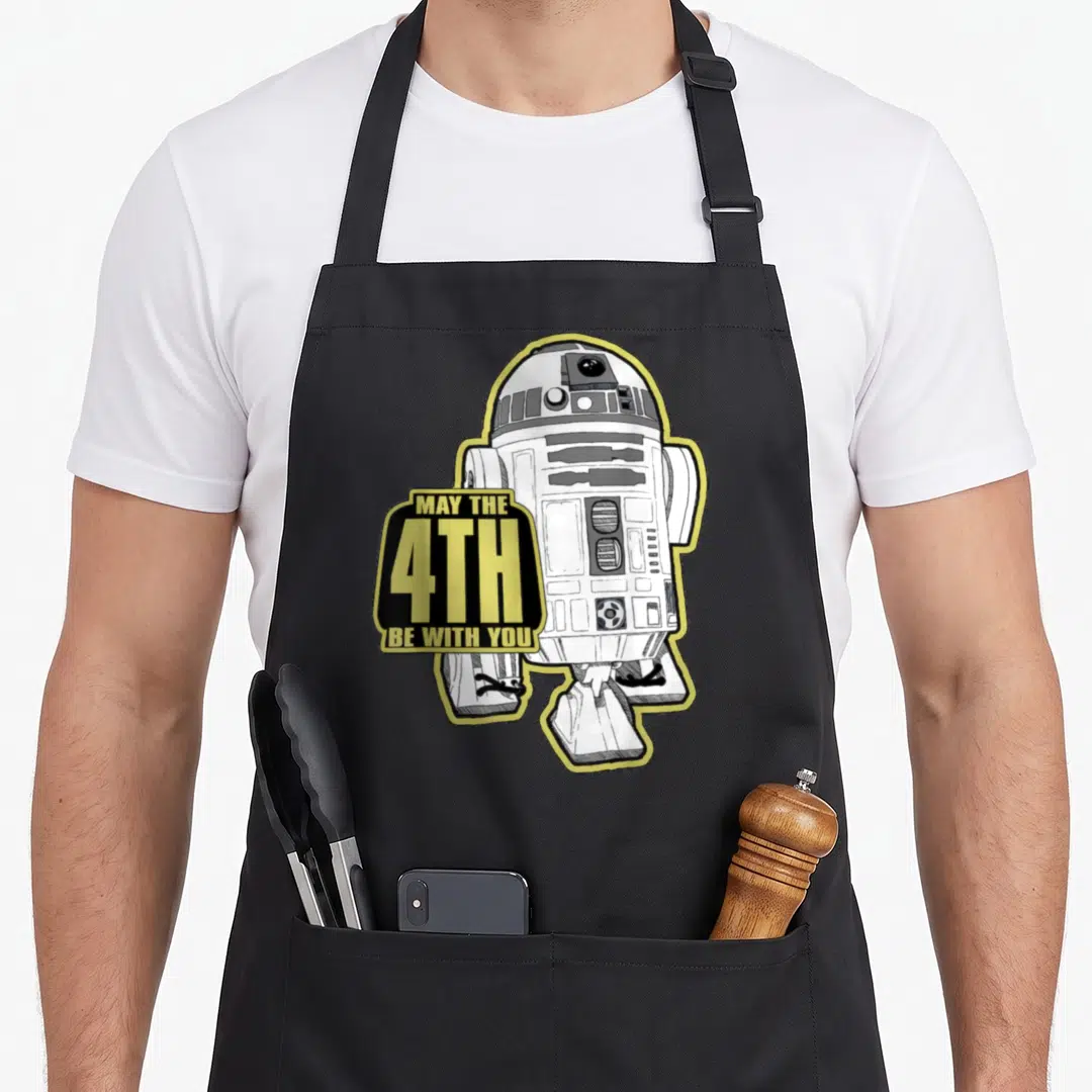 May worlds greatest apron - R2-D2 robot with yellow sign saying MAY THE 4TH BE WITH YOU in cartoon style — shown on a black cotton canvas apron against a white background