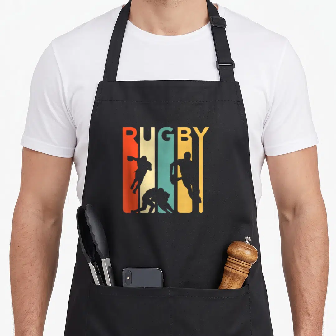 Rugby 1970s apron - Rugby text in red cream teal yellow orange stripes with three grey player silhouettes — shown on a black cotton canvas apron against a white background