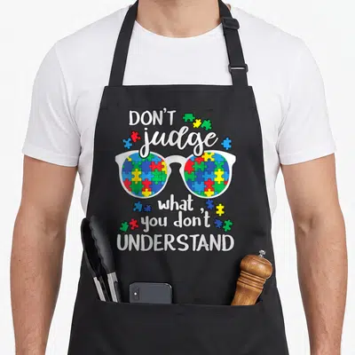 Judge walking apron - Glasses with colorful puzzle piece lenses surrounded by text reading DON'T judge what you don't — shown on a black cotton canvas apron against a white background