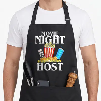 Night outfit family aprons - Cartoon popcorn bucket, tickets, remote, and drink between white MOVIE NIGHT HOST text — shown on a black cotton canvas apron against a white background