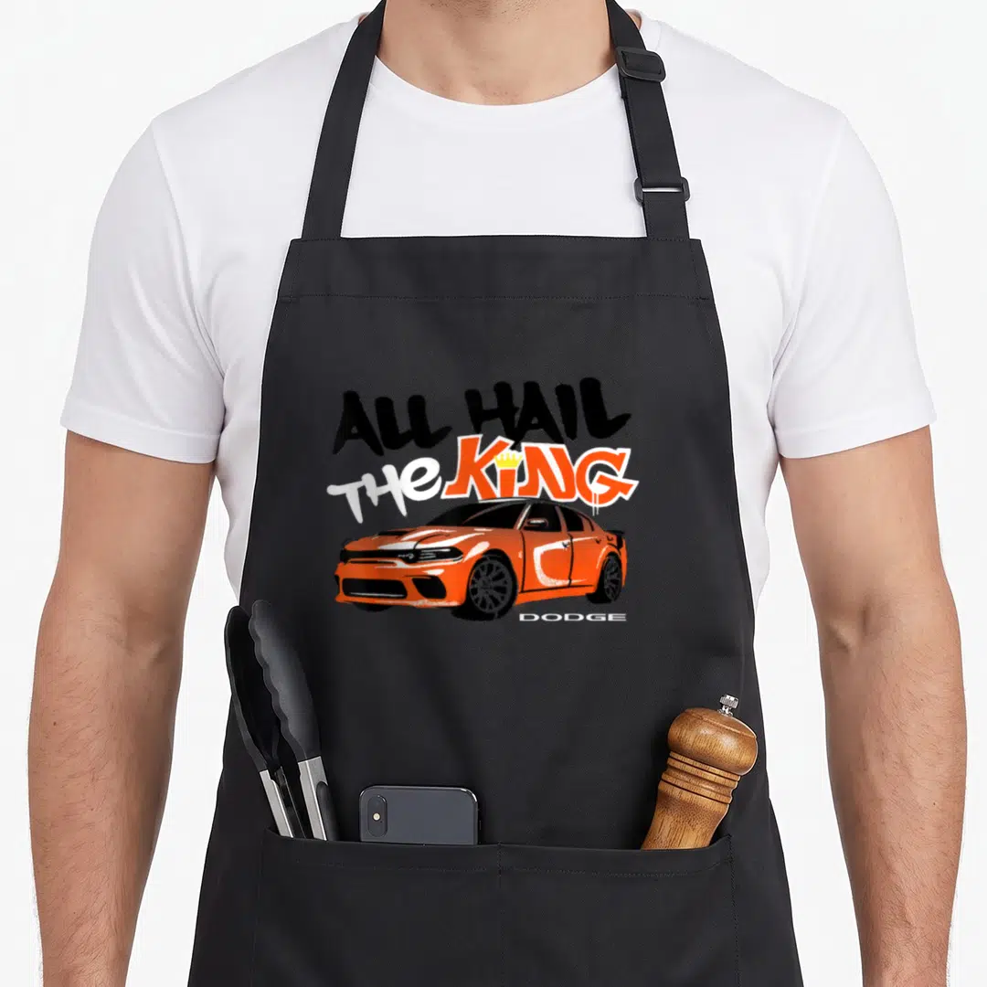 Dodge king apron - Orange cartoon Dodge sedan with All Hail The King text and small yellow crown — shown on a black cotton canvas apron against a white background