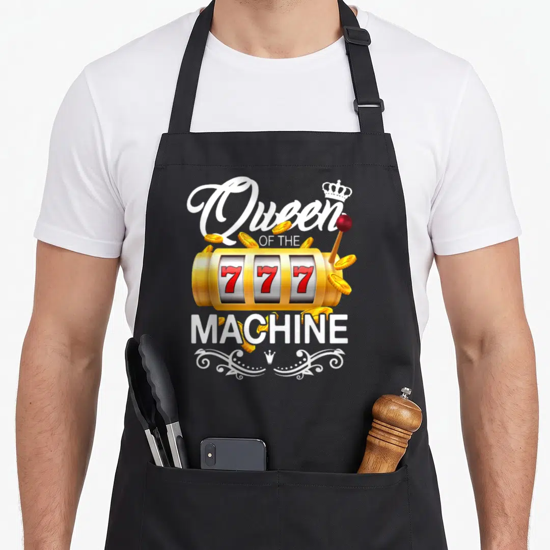 Queen machine apron - Gold slot machine with red 777 reels and white Queen OF THE MACHINE text — shown on a black cotton canvas apron against a white background