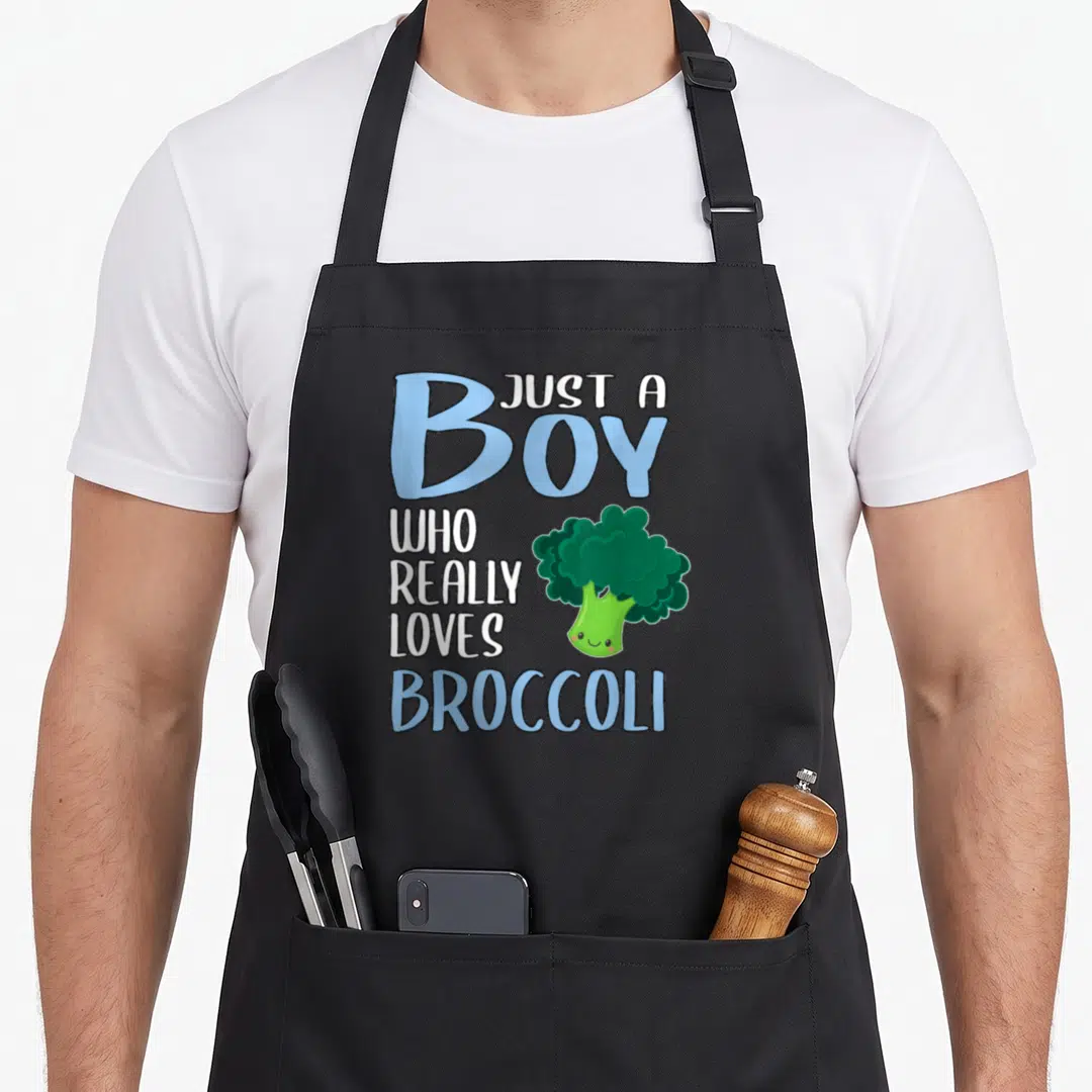 Cute broccoli apron - Just A Boy Who Really Loves Broccoli text with smiling cartoon vegetable in blue and green — shown on a black cotton canvas apron against a white background